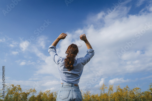 Back view of a woman standing outdoors with arms raised toward a blue sky and clouds, symbolizing freedom, happiness, success, inspiration, confidence, wellness, hope and a positive lifestyle.