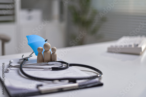 Fotografie Wooden family figures under blue umbrella with stethoscope on medical clipboard
