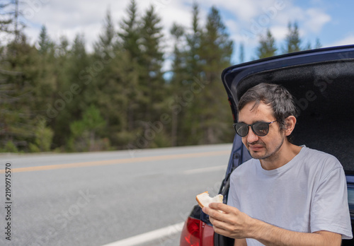 A traveller has a snack while sitting near a car