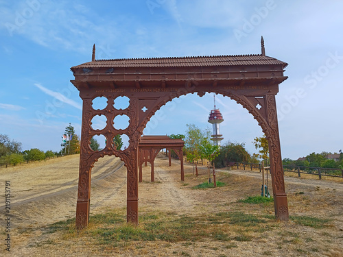 Foto The Székely gates on the Vértói Hill in Szeged, one after another, photographed from the bottom of the hill, in the sunlight, with leafy trees, and behind them Szeged's iconic telephone tower