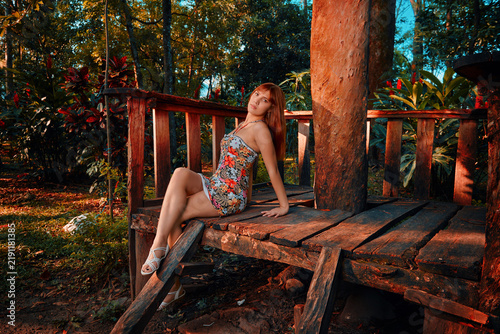 Fotografie A beautiful young Latina with reddish-brown hair sits cross-legged on a wooden kiosk floor, her flowery dress glowing in sunset light