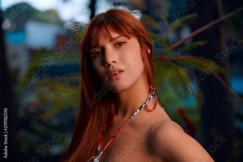 A close-up of a beautiful young Latina with reddish-brown hair in a flowery dress at a park, her face glowing in sunset light.