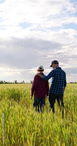 Farmer father walks through green wheat field with son, talking and smiling. Father keeps arm around boys shoulders, showing close family bond, rural lifestyle, care and connection with nature.