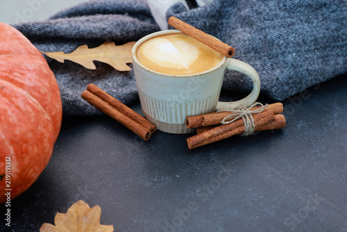 Aromatic spicy coffee with milk and cinnamon sticks at gray scarf and dry leaves on black table. Cup of latte close up. Autumn menu, hot seasonal drink, thanksgiving time