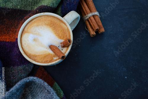 Cup of spicy latte and warm woolen scarf on dark background with copy space. Hot coffee with evaporated milk and cinnamon sticks. Winter menu concept, warming drink