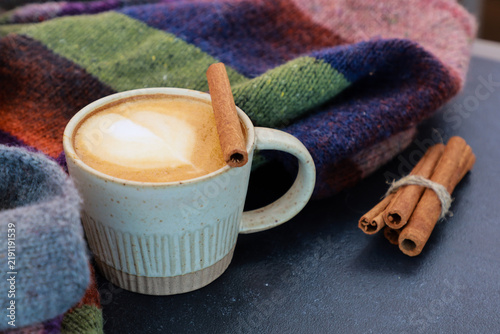 Close up of spicy latte with cinnamon stick at knitted scarf on black table. Cozy warming coffee break, winter morning, coffee shop menu concept