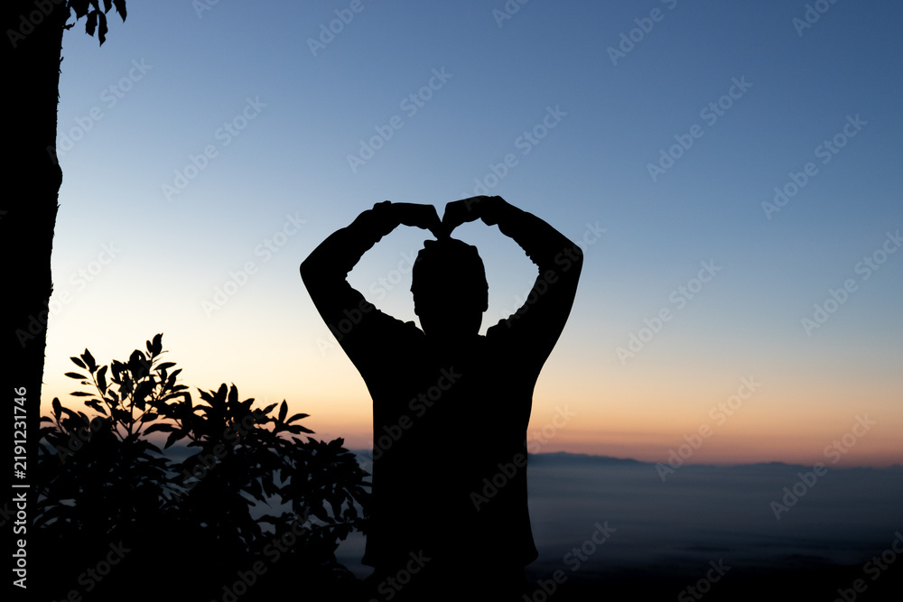 Obraz premium Silhouette of person forming heart gesture with hands against morning sunrise sky.