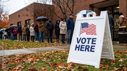 A white sign with an American flag and VOTE HERE text stands near people waiting to vote.