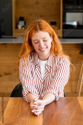 Картината върху платно Redhead woman smiling and relaxed at a wooden kitchen table in a striped shirt, eyes closed reflecting contentment and calm, lifestyle portrait of comfort during quiet home moment