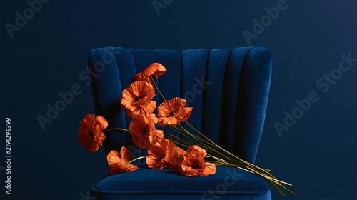 Blue velvet armchair with a bunch of orange poppies on it. the armchair has a curved backrest and armrests, and the background is a solid dark blue color.