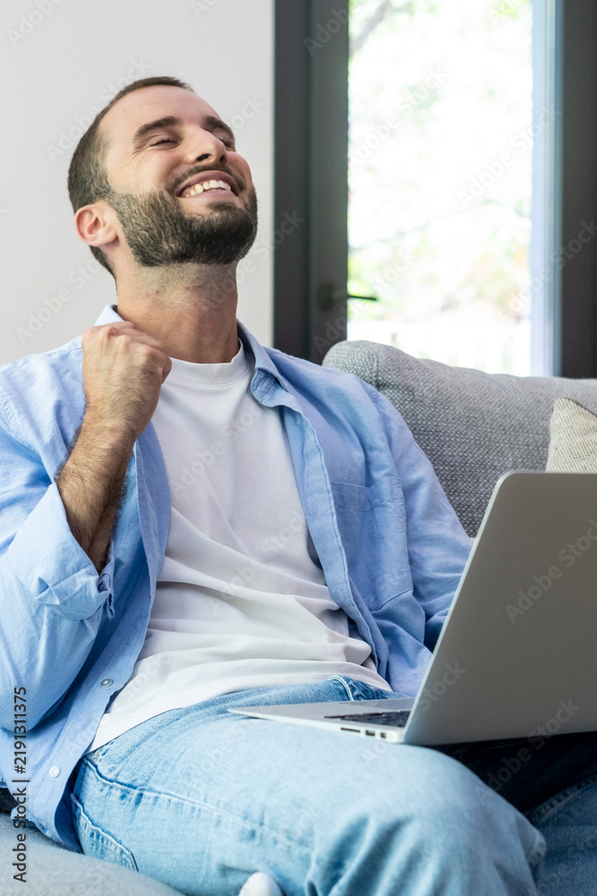Fototapeta premium Man celebrating success working on a laptop at home, relaxed on a couch with a joyful expression, remote work achievement and confidence in a bright, comfortable living room.