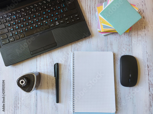 Top view of a black laptop, computer mouse, open notebook with a pen, and notepaper on a light wooden background. Workspace with office supplies for work, freelance and online study