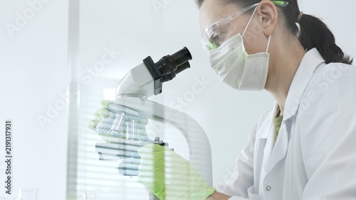 Fotografie Female scientist wearing protective mask and safety goggles looking through a mi