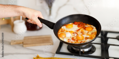 Quadro su tela Hand holding skillet with eggs in spicy tomato sauce over gas burner for breakfa