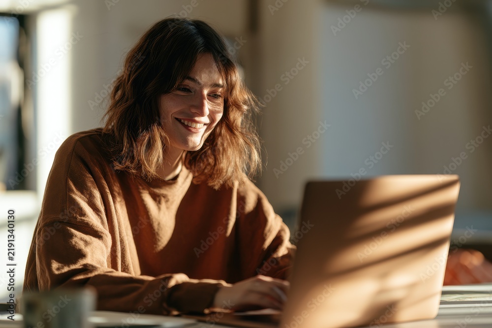 Obraz premium Joyful woman smiles while using a laptop in a bright home office setting