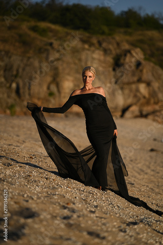 A beautiful blonde woman in a long black dress stands on the beach. Sunset.