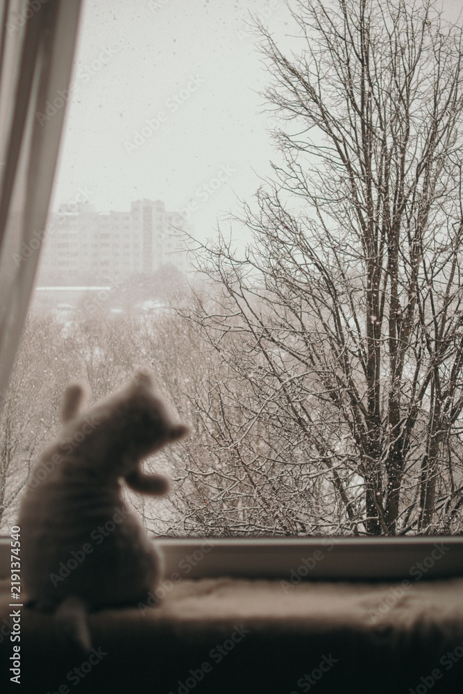 Fototapeta premium Plush cat toy on a windowsill against snow-covered trees and distant apartment buildings during winter snowfall.