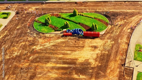 Aerial view of a construction site with a landscaped roundabout, green grass, small trees, bare earth, and construction materials including blue pipes and red bundles in Latvia.