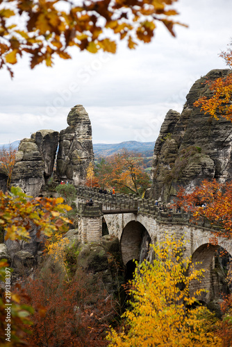 Obraz na płótnie Vertical shot of the iconic sandstone Bastei Bridge with tourists framed by warm yellow and orange autumn foliage in Saxon Switzerland