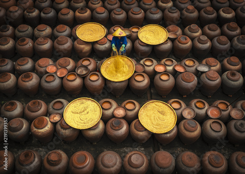 Aerial view of a worker wearing a conical hat sorting yellow grains on trays amidst rows of large ceramic jars at a soy sauce factory in Ban Yen Nhan, Vietnam.