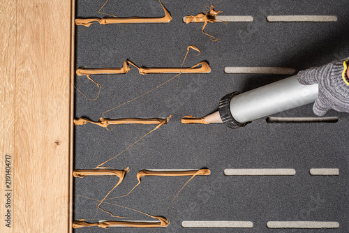 Worker applying adhesive glue with applicator gun onto acoustic underlayment for parquet installation