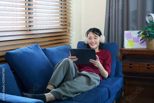 Young happy woman lying in living room relaxing on sofa listening to music in headphones using tablet at home, enjoying leisure time and online, modern relaxation and digital engagement