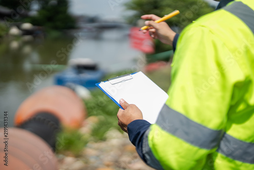Male environmental engineer records field observations on clipboard while checking water conditions and drainage infrastructure during environmental site assessment.