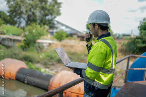 Male environmental engineer communicates by radio while using laptop during environmental monitoring and water infrastructure assessment at drainage facility.