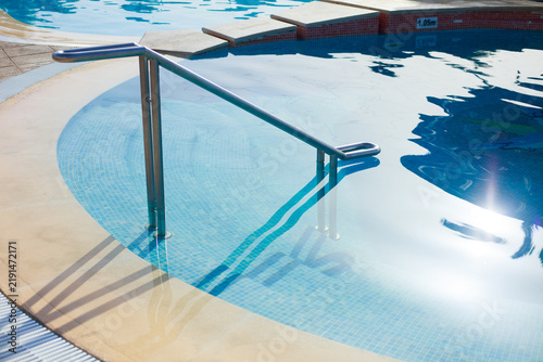 Outdoor swimming pool with stainless steel handrails, shallow tiled steps, clear blue water and bright sunlight reflecting across the pool surface.