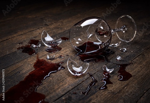 A shattered wine glass with spilled red wine on a rustic dark wooden surface representing accident, loss, or celebration aftermath