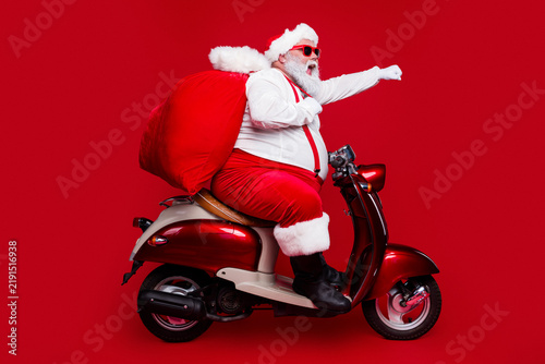 Fotografie Santa Claus on a red scooter with gift sack striking a playful superhero pose —