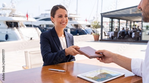 Smiling woman in business suit handing leather notebook to man at table in yacht marina. Luxury harbor with boats and restaurant terrace in background.
