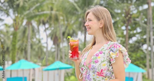Happy young woman holding cold tropical cocktail at luxury swimming pool resort with palm trees. Smiling female tourist enjoying refreshing beverage on summer vacation.