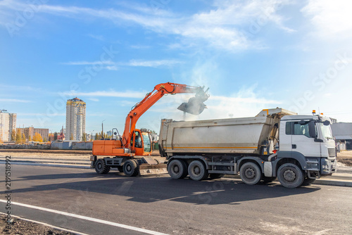 Quadro su tela Orange wheeled excavator loading earth material into a heavy white dump truck on a new asphalt road during infrastructure construction