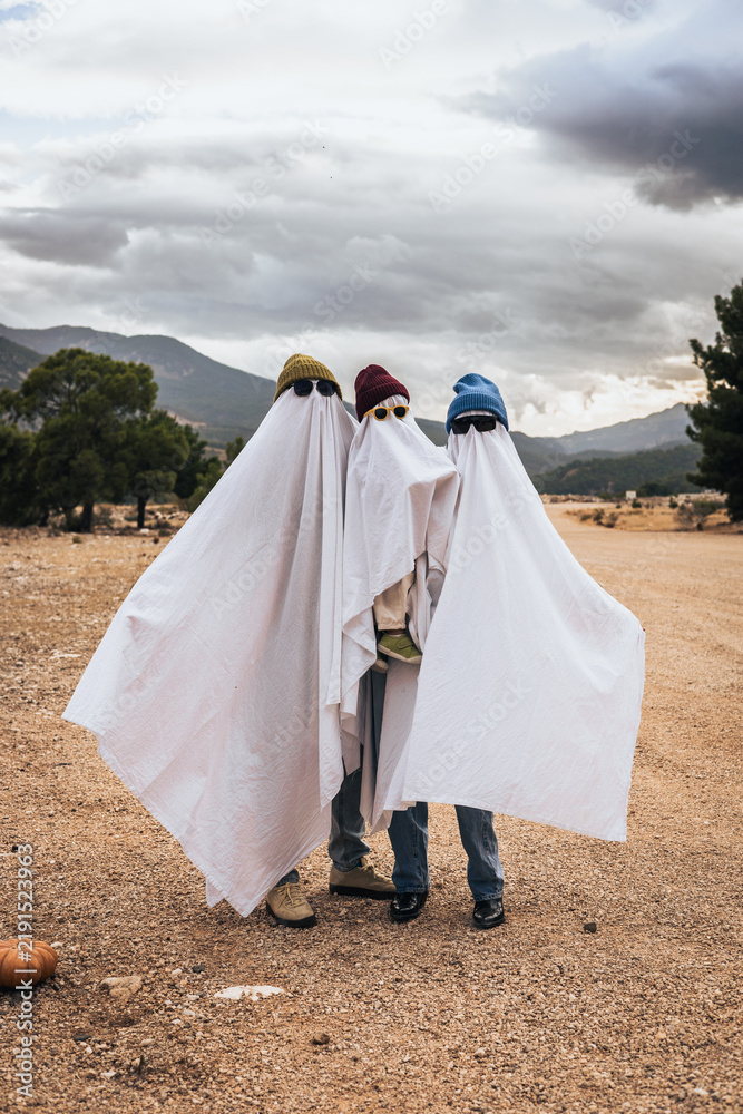 Fototapeta premium Three children dressed as ghosts for Halloween, wearing white sheets and hats. They stand together in a rural area with mountains in the background.