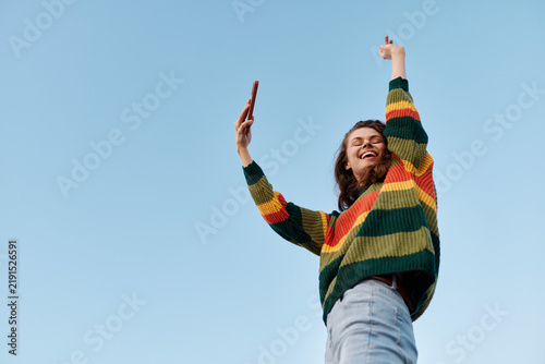 Quadro su tela Joyful woman stands outdoors, wearing a vibrant striped sweater, arms raised in celebration, against a clear blue sky