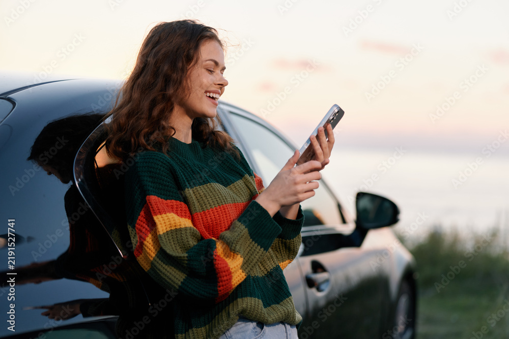 Obraz premium Smiling young woman in a colorful striped sweater leans on a car and uses her smartphone at sunset by the sea. Travel and leisure concept.