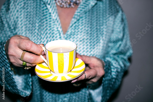 Woman holding a yellow and white striped teacup with saucer, wearing a turquoise patterned blouse and silver jewelry, against a neutral background