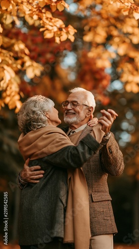 an elderly couple embracing in a park during autumn. The man, wearing glasses and a checkered blazer, holds the woman close. The woman, with curly silver hair, wears a dark coat and a beige scarf.
