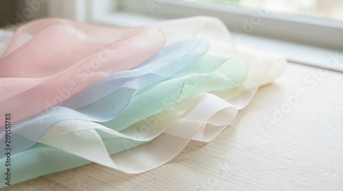 Layered pastel organza fabric on wooden table. Transparent chiffon silk cloth folds near window