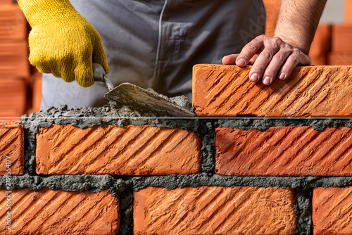 Close-up shot of a bricklayer laying red clay bricks with mortar using a trowel, representing bricklaying work, wall construction, masonry building site, and professional contractor labor.