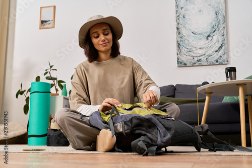 Canvastavla Young woman in safari hat prepares her hiking backpack in a stylish living room