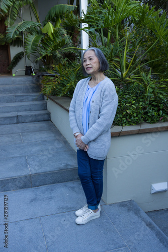Senior Asian woman standing on tiled walkway at home entrance by raised planter and stone steps