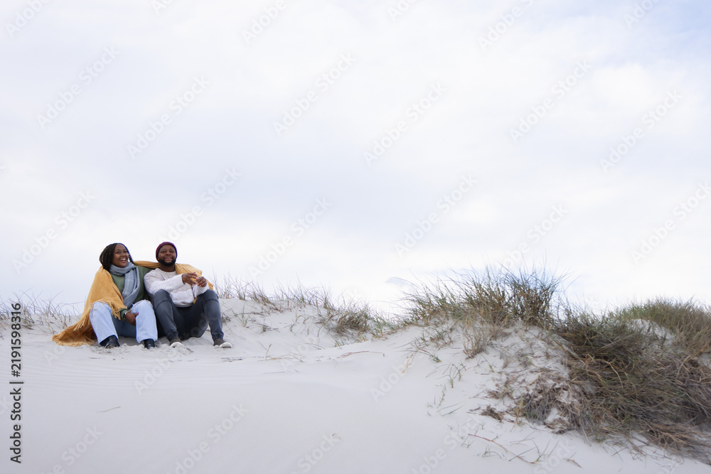 Obraz premium Couple sitting on sand dune at beach wrapped in yellow blanket wearing burgundy beanie, copy space