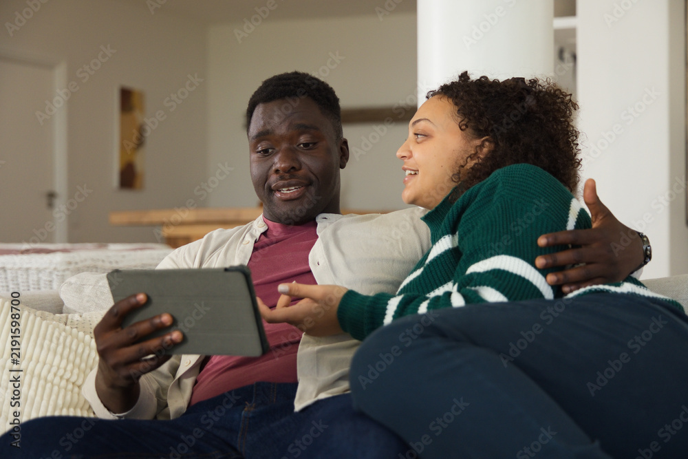 Obraz premium Couple sitting closely on light-colored sofa in living room, holding tablet and pointing at screen
