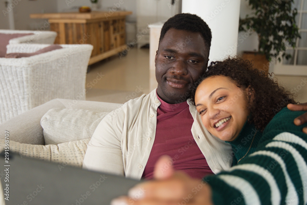 Obraz premium African American couple sitting on living room sofa, leaning and smiling while watching smartphone