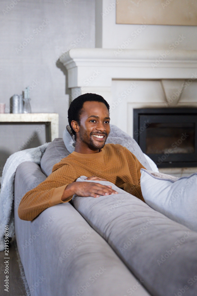 Obraz premium African American man reclining on gray sectional sofa in cozy living room, smiling in brown sweater