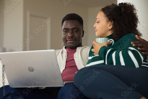 African American couple sitting on light sofa at home, sharing silver laptop and light blue mug