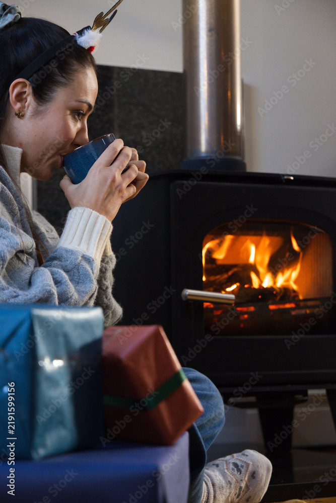Obraz premium Female sitting by wood-burning stove in living room sweater sipping blue mug near gifts, copy space