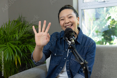 Asian woman sitting on grey sofa wearing denim shirt, waving hand at microphone, smiling